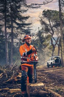 A person in work gear holding a chainsaw in a forest with cut logs and a vehicle in the background.