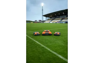 Three robotic lawn mowers on a football field, with stadium seating in the background.