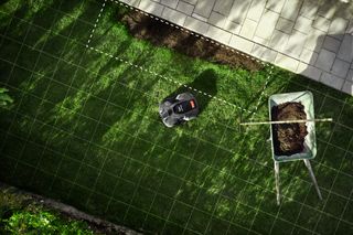 Robotic lawn mower operating on a grass lawn with grid lines overlay, next to a wheelbarrow with soil.
