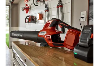A Husqvarna leaf blower and battery charger on a wooden table in a garage setting.