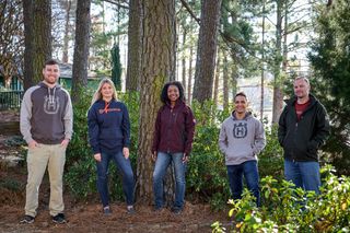 Five people standing in a forested area, wearing casual and work-related clothing.