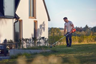 A person using a grass trimmer to maintain the lawn near a modern house.