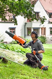 A person kneeling on grass using a leaf blower in a garden with a house in the background.