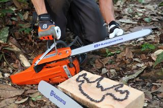 A person kneeling in a forest setting, adjusting a Husqvarna chainsaw next to a wooden block with a chainsaw chain.