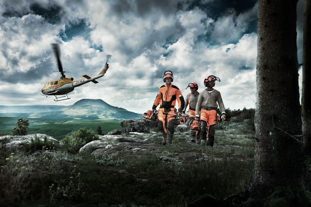 Three workers in work gear carrying chainsaws, with a helicopter flying over a mountainous landscape.