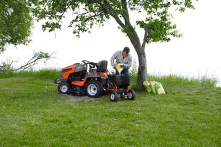 A person loading a ride-on mower with gardening supplies under a tree in a grassy area.