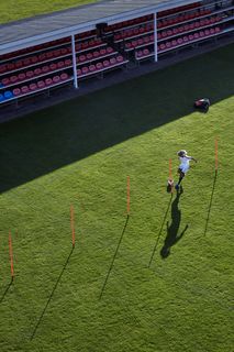 A person running on a grassy field with training poles set up, near a stadium seating area.