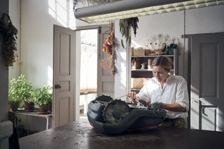A person working on a robotic lawn mower in a workshop setting, surrounded by plants and tools.
