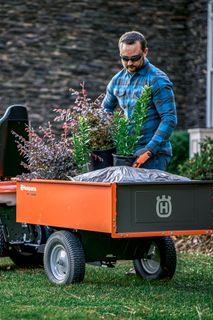 A person loading plants and a bag into a Husqvarna ride-on mower trailer in a garden.