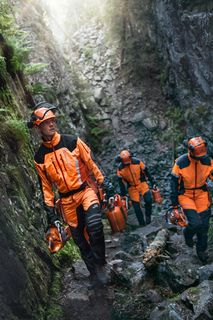 Three workers in rugged terrain carrying construction equipment, wearing work gear.