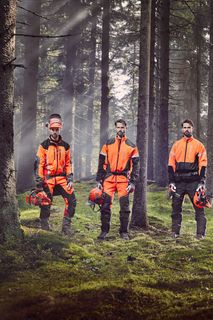 Four people in work gear standing in a forest holding helmets and chainsaws.