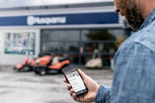 A person holding a smartphone displaying the Husqvarna Connect app, with Husqvarna products in the background.