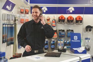 A person in a store setting, standing at a counter with Husqvarna safety helmets and products displayed on the wall.