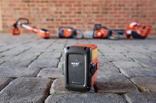 Close-up of a Husqvarna battery on a cobblestone surface with various Husqvarna tools in the background.