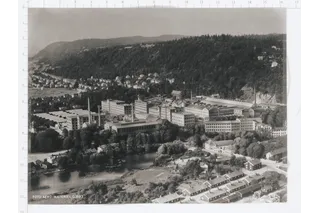 Aerial view of a large industrial complex surrounded by forested hills and a river.
