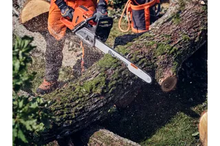 A person using a Husqvarna chainsaw to cut through a moss-covered tree log outdoors.