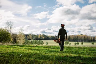 A person walking across a grassy field carrying a chainsaw, with a forest in the background.