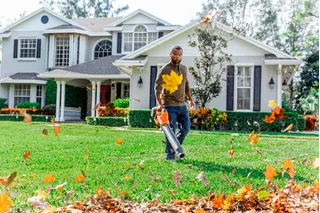 A person using a leaf blower to clear fallen leaves from a pathway in front of a large house.