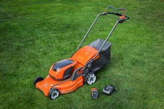 Husqvarna battery-powered lawn mower on a lawn with a battery and charger beside it.