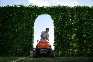 A person riding a lawn mower through an archway in a tall hedge.