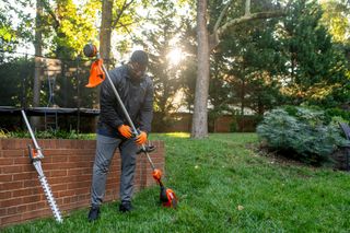 A person adjusting a garden trimmer in a backyard setting, with a hedge trimmer resting nearby.