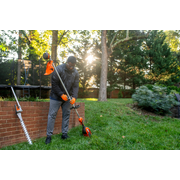 A person adjusting a garden trimmer in a backyard setting, with a hedge trimmer resting nearby.