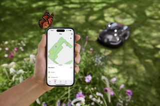 A hand holding a smartphone displaying a garden map app, with a robotic lawn mower operating in the background and a butterfly perched on the phone.