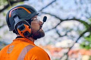 A person wearing work gear and a helmet with ear protection stands in focus, while another person is blurred in the background standing on a tree branch.