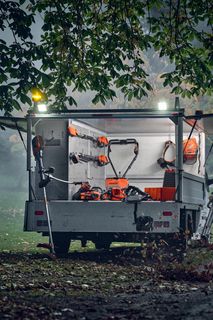 A person using a leaf blower to clear fallen leaves in a park setting, with a trailer containing various gardening tools.