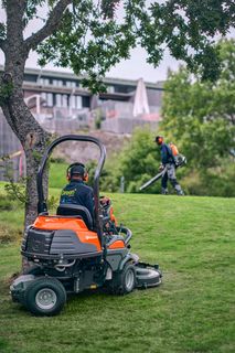 A person mowing a lawn with a ride-on mower while another person uses a trimmer in a garden.