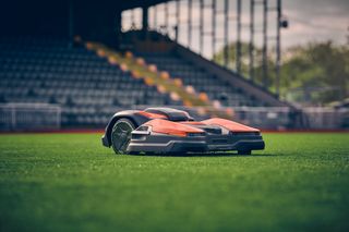 Robotic lawn mower operating on a sports field with empty stands in the background.